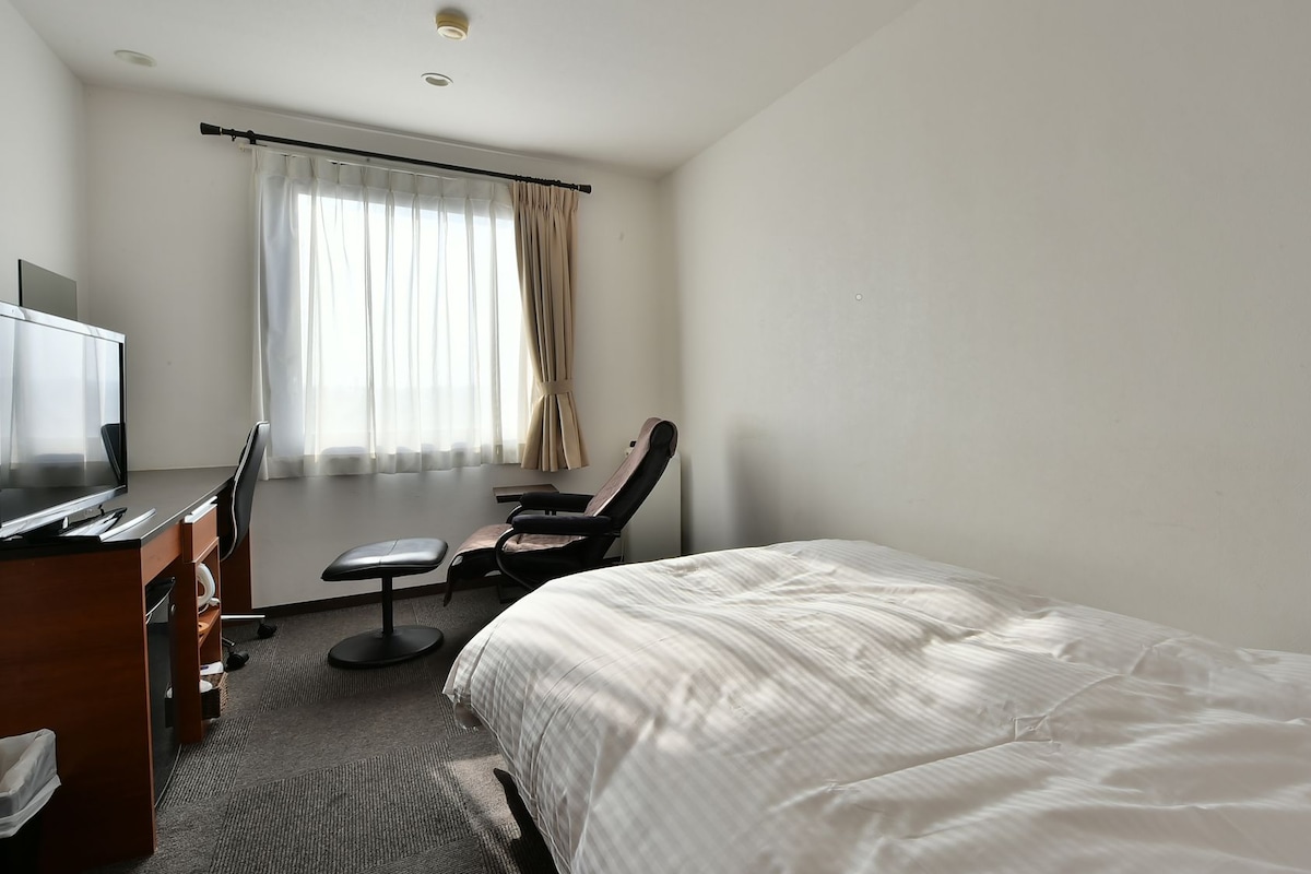 A hotel room with a bed, a desk with a chair, a TV, and a window with curtains.