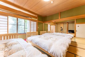 Une pièce de style japonais traditionnel, avec un sol recouvert de tatamis, un grand lit avec une literie blanche, un téléviseur posé sur un meuble en bois et des portes coulissantes donnant sur une autre pièce.