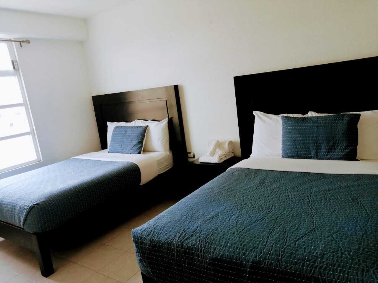 Dos camas individuales con cabeceros oscuros y almohadas a juego, una mesita de noche con teléfono y una ventana con cortinas.