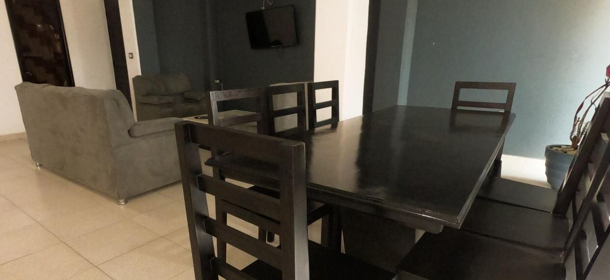 A dining area with a dark wooden table and chairs, a grey sofa, and a flat-screen TV mounted on the wall.