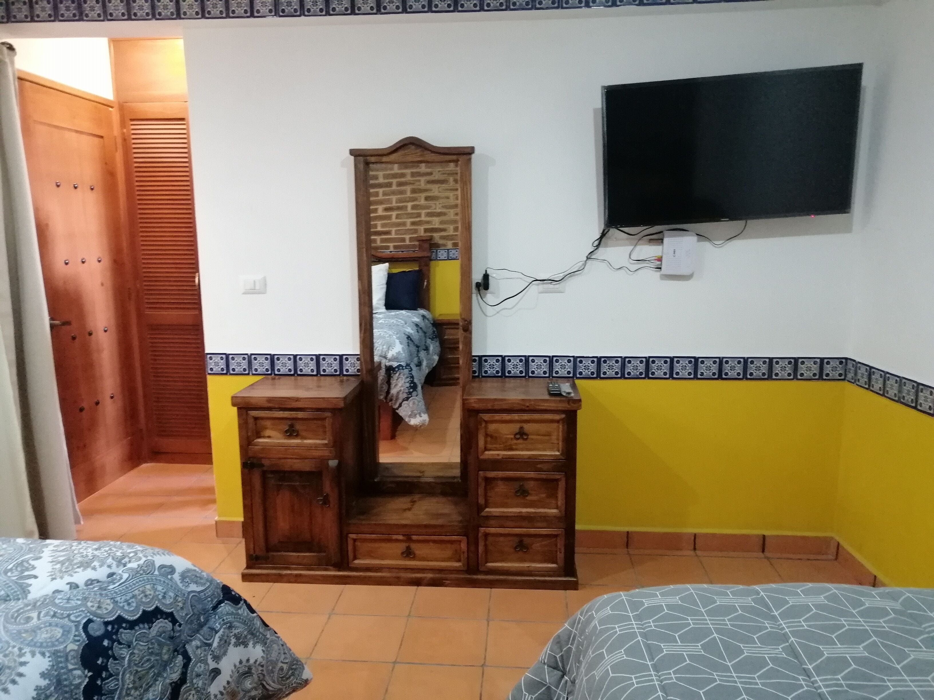 A bedroom with a wooden dresser, a bed with patterned bedding, a mirror, and a television mounted on the wall.