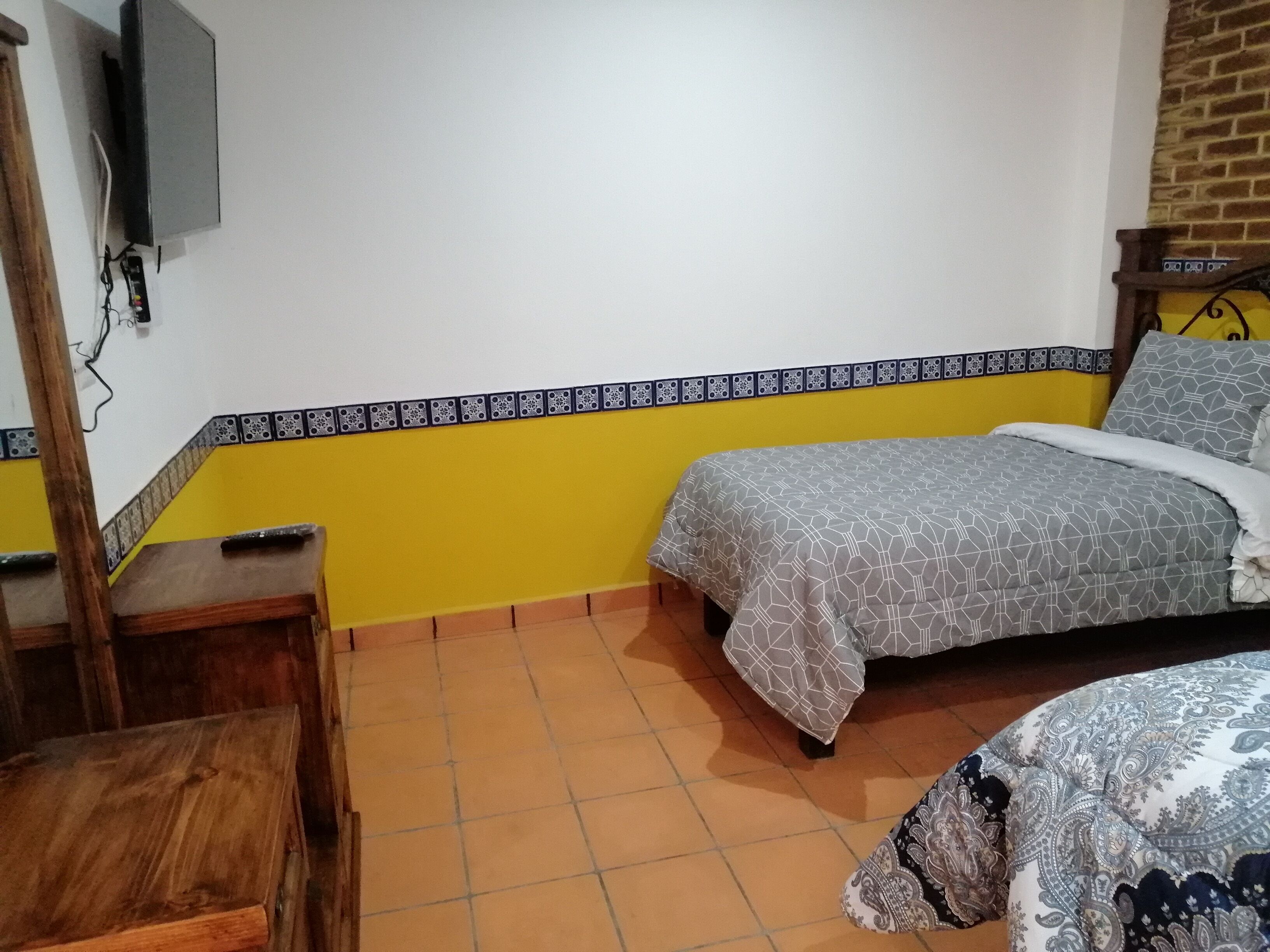 Un dormitorio con una cama, una mesita de noche de madera, un televisor montado en la pared y piso de baldosas.