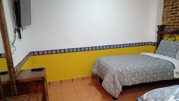 Un dormitorio con una cama, una mesita de noche de madera, un televisor montado en la pared y piso de baldosas.