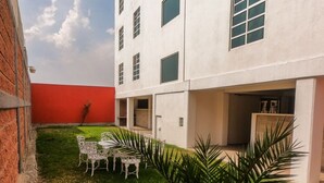 A courtyard with a white building, a red wall, and a small lawn with a table and chairs. - Hotel y Suites Axolotl (Chignahuapan)