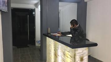 Un mostrador de recepción con una persona detrás del mostrador, una puerta al fondo y suelo de baldosas.