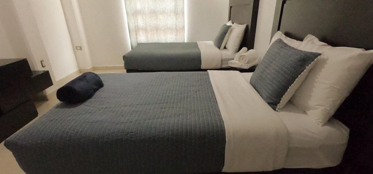 A double bed with a grey and white duvet, a black pillow, and a grey pillow. There is a small bedside table with a lamp.