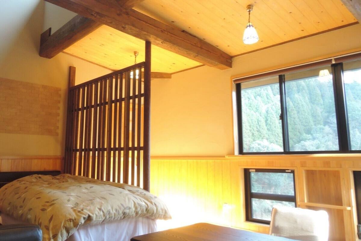 A room with a bed, a wooden partition, a window with a view of trees, and a lamp on a bedside table.