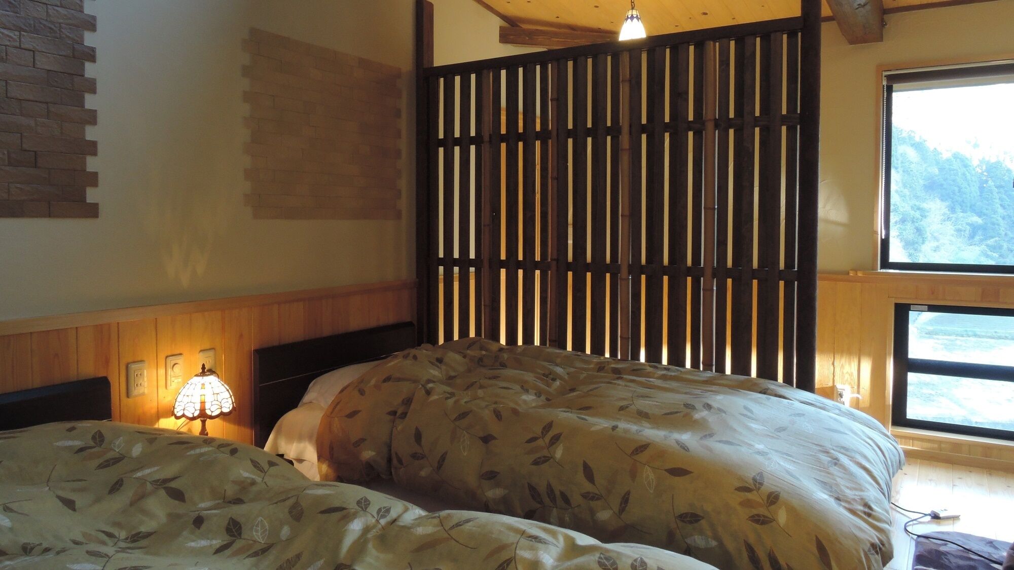 Una camera da letto con due letti, una testiera in legno, una lampada a parete e una finestra con vista sugli alberi.