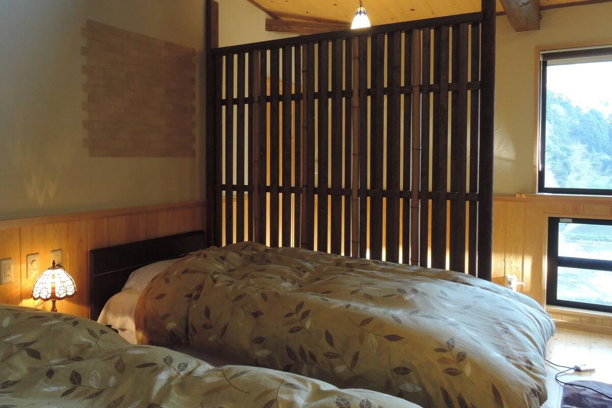 A bedroom with two beds, a wooden headboard, a wall-mounted light, and a window with a view of trees.