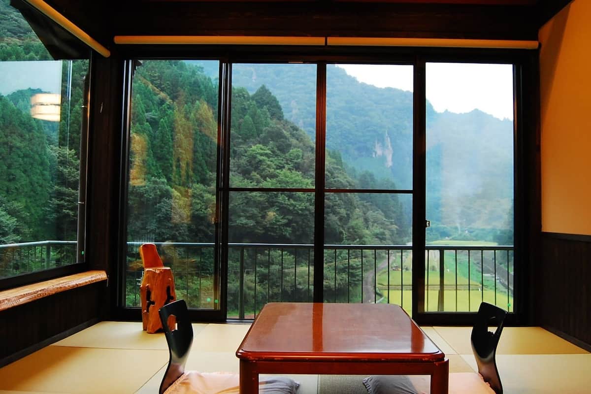 A room with tatami flooring, a wooden table, and a view of a mountainous landscape through large windows.