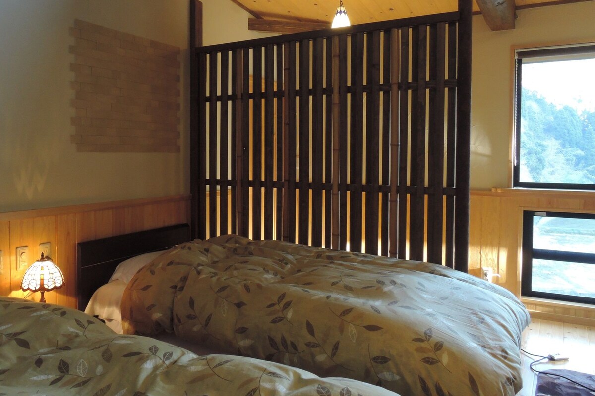 A bedroom with two beds, a wooden headboard, a wall-mounted light, and a window with a view of trees.