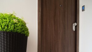 Une porte en bois sombre, dotée d’une petite serrure électronique, un grand pot de fleurs placé à gauche, et un paillasson noir avec des lettres dorées sur le sol carrelé.