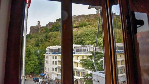 A view through a window with a metal frame, featuring a building with multiple stories, a balcony, and a hill with a structure on top. - Hotel Sofra (Gjirokastër)