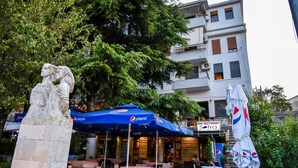 An outdoor seating area with tables and umbrellas, a stone statue, and a building with balconies and signage. - Hotel Sofra (Gjirokastër)