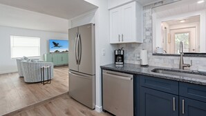 Una cocina moderna con un refrigerador de acero inoxidable, lavavajillas y gabinetes azul oscuro. Hay una ventana con persianas y un cuadro de un pájaro en la pared.