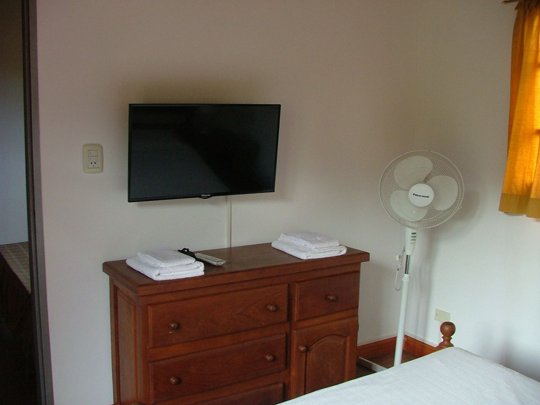 A hotel room with a wooden dresser, a flat-screen TV mounted on the wall, a standing fan, and a bed with a white comforter.
