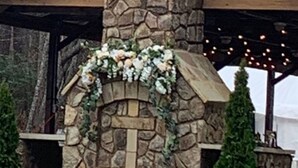 Una chimenea de piedra con un arreglo floral, rodeada de sillas blancas y follaje.