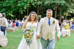 A wedding couple walking hand in hand, surrounded by guests and greenery.