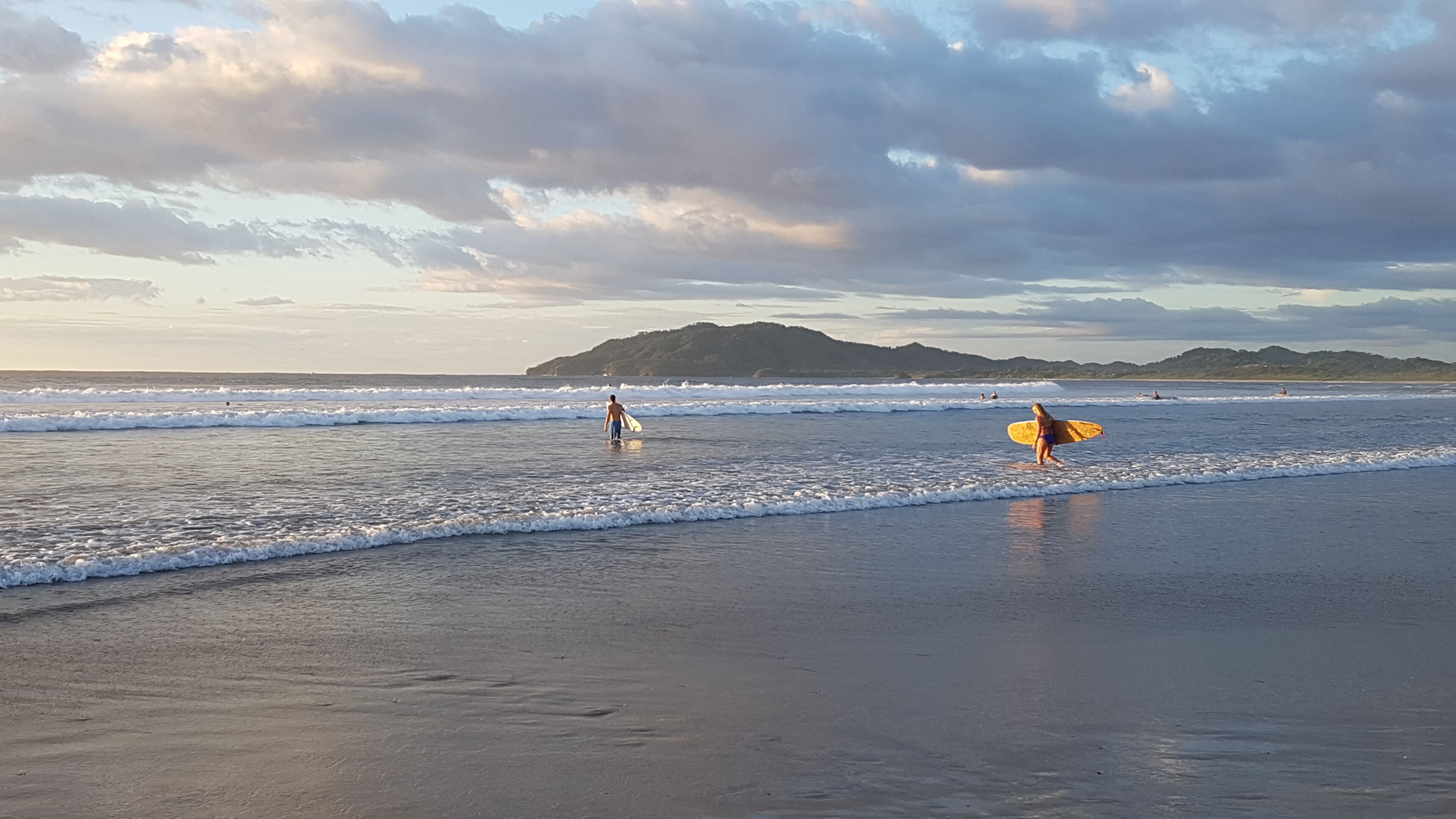 A beach with waves, a distant mountain, and a person with a surfboard.