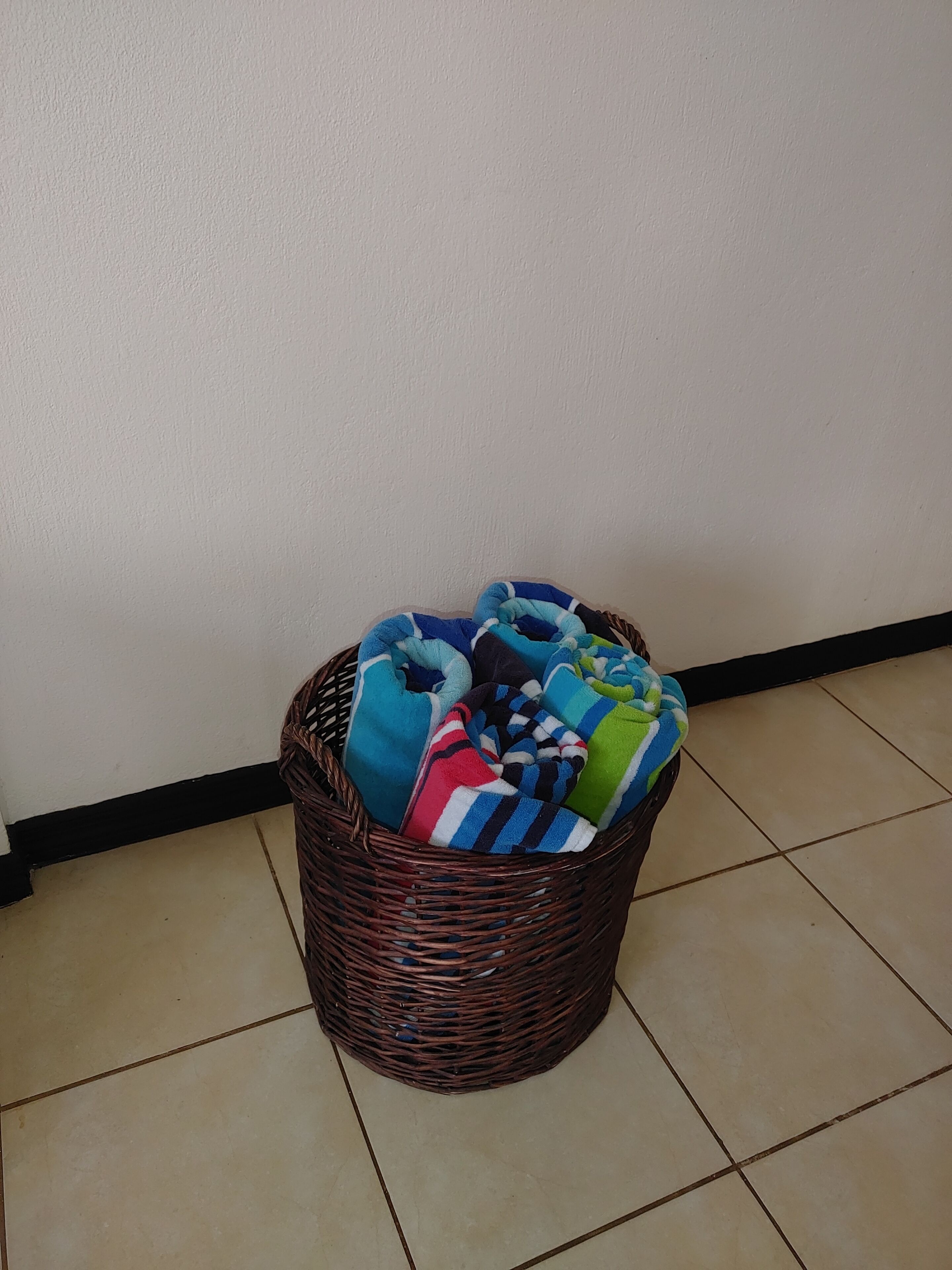 Un panier en osier rempli de serviettes colorées, posé sur un sol carrelé devant un mur uni.