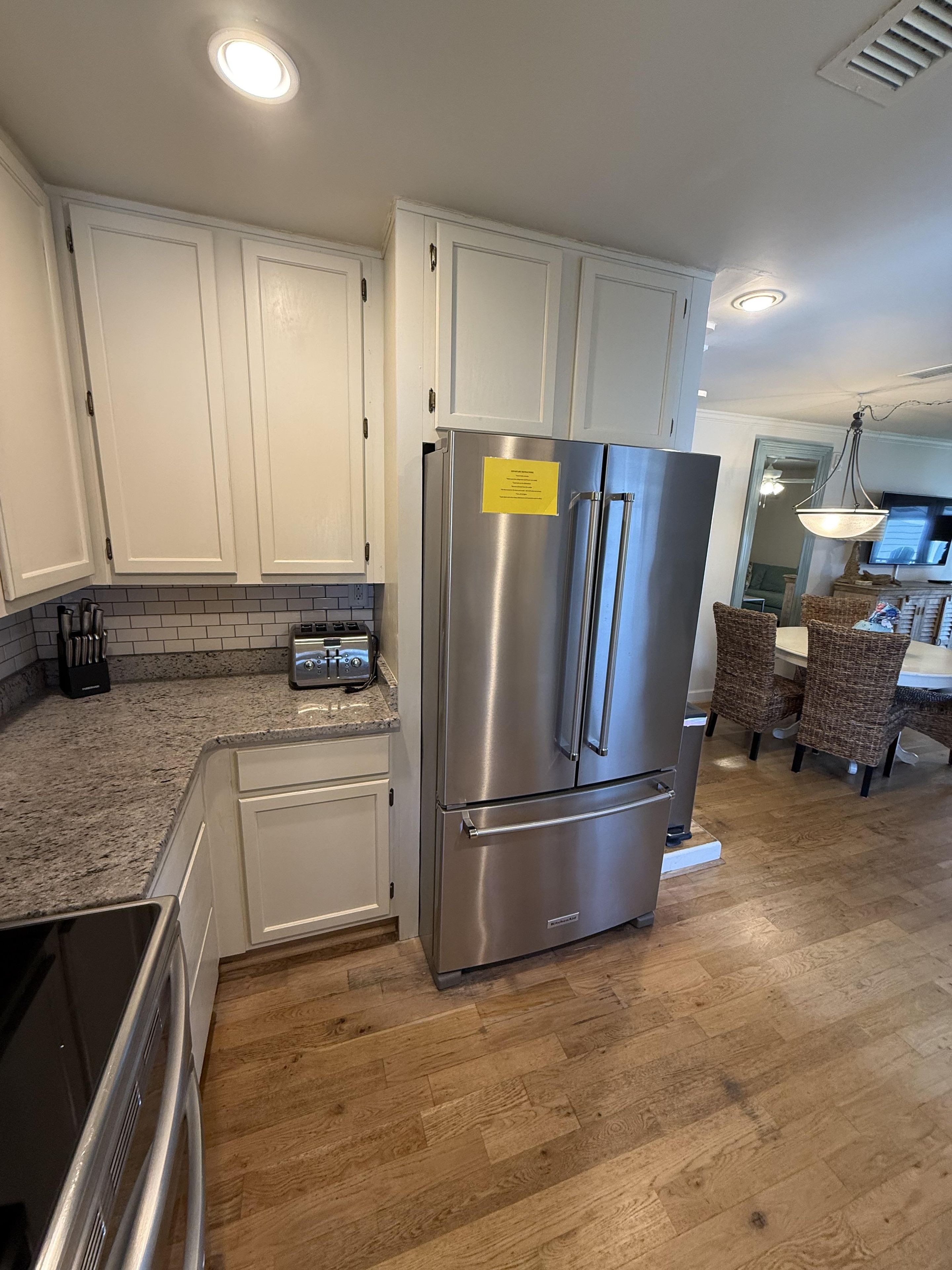 A kitchen with white cabinets, a stainless steel refrigerator, a granite countertop, and a dining area with wicker chairs.