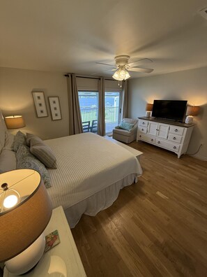 A bedroom with a bed, a dresser, a television, and a view of the outdoors. - C21 Sundown Riverside Condominiums (Pensacola)