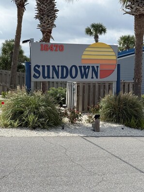 A sign with the name 'SUNDOWN' and the address '16470' in a landscaped area with palm trees and shrubs. - C21 Sundown Riverside Condominiums (Pensacola)