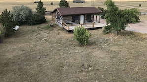 A rustic log cabin with a wooden deck, surrounded by sparse vegetation and a vast open field.