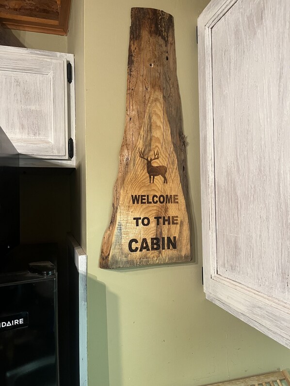Uma placa de madeira com silhueta de veado e o texto 'WELCOME TO THE CABIN' fixada em uma parede.