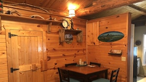 A wooden interior with a dining table and chairs, a clock, and a landscape painting.