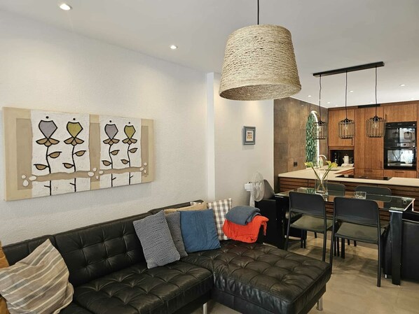 A modern living room with a black leather sofa, a dining area with chairs, a kitchen with wooden cabinets, and a hanging woven light fixture.