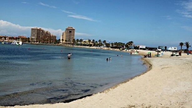 Ein Strand mit blauem Himmel, sandigem Ufer und ein paar badenden Personen. Im Hintergrund sind Gebäude zu sehen, darunter ein hohes Hotel.