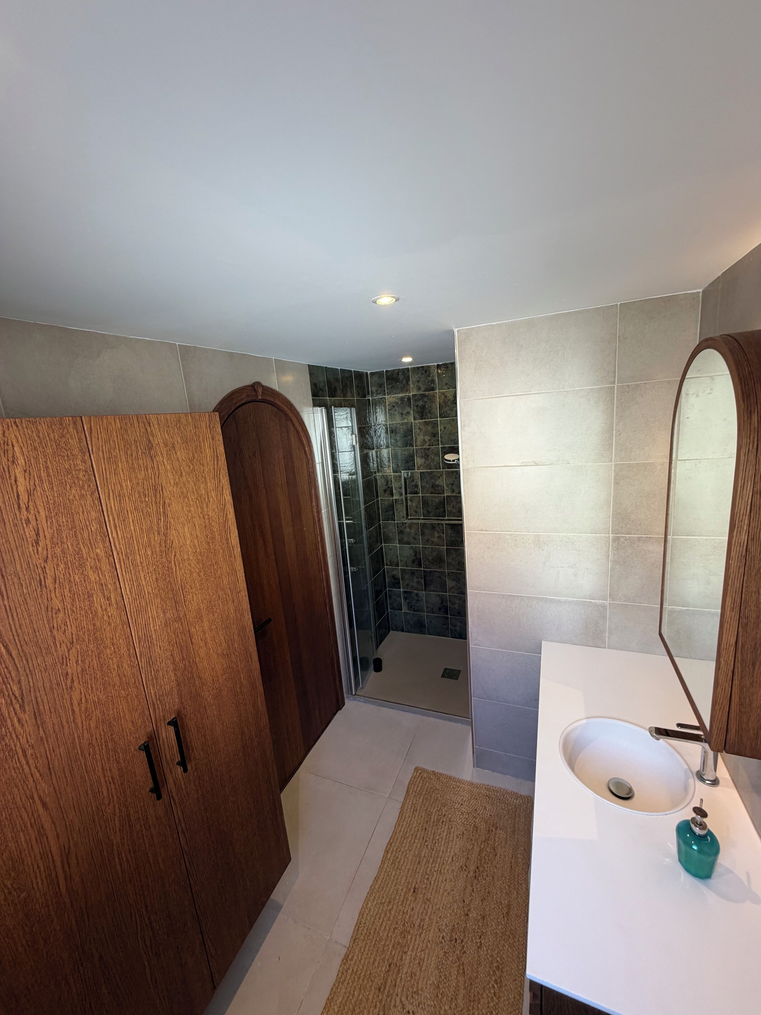 A bathroom with a wooden door, a glass shower enclosure, a white sink with a mirror, and a beige rug.