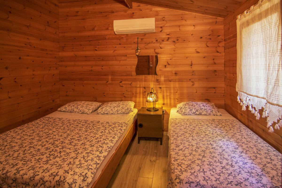 A wooden cabin room with two beds, a nightstand, an air conditioning unit, and a window with a curtain.