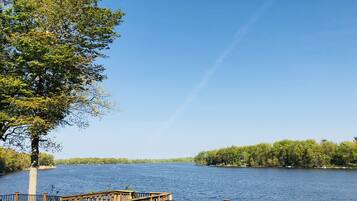 Una terraza de madera con baranda metálica que da a un lago tranquilo, rodeado de árboles y un cielo despejado.