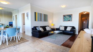 A modern living room with a black leather sofa, a coffee table, and a dining area with a white table and blue chairs. - Breathtaking Ocean Views - 2 bedroom apartment with parking (Tamarama)