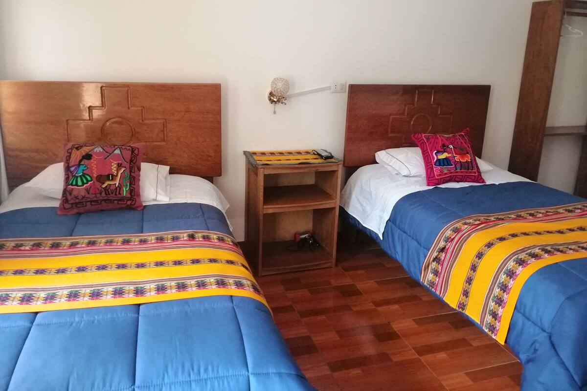 Two beds with blue bedding and colorful pillows, a wooden nightstand with a lamp, and a wooden wardrobe.