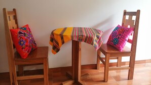 Dos sillas de madera con almohadones coloridos, una pequeña mesa de madera con mantel estampado y una pared blanca lisa.