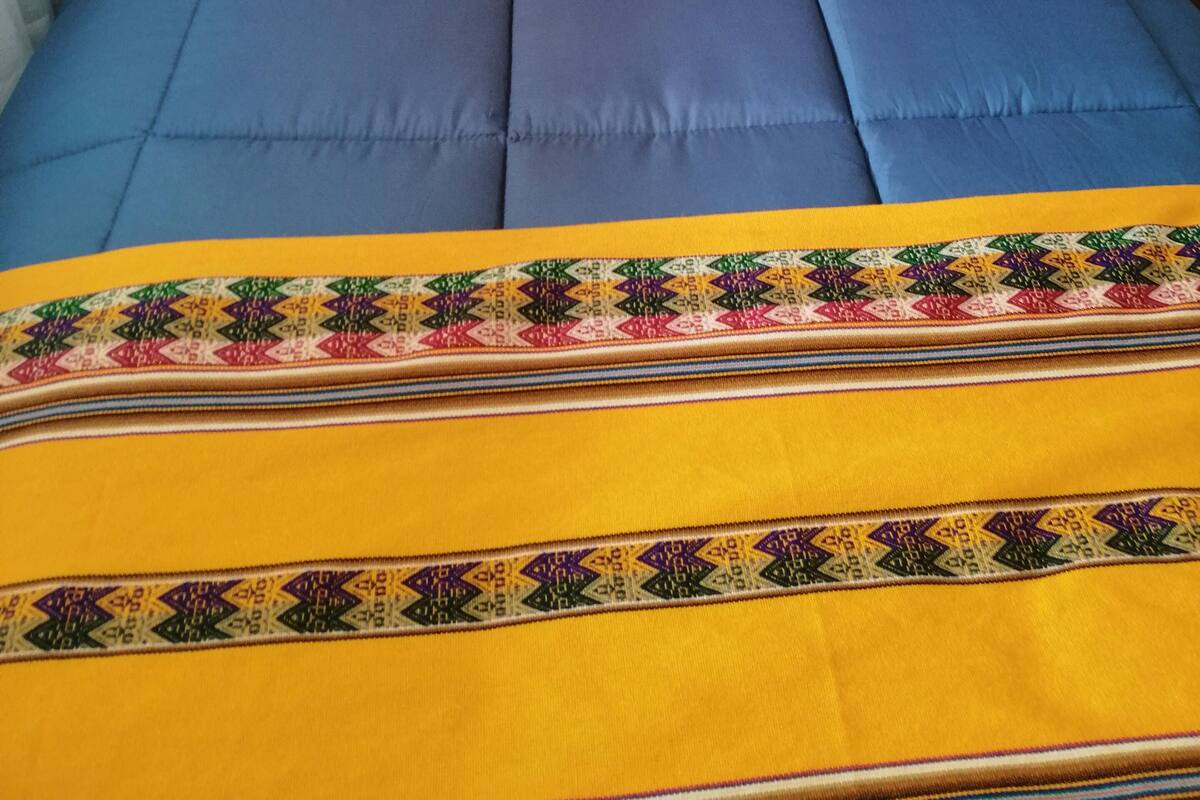 A bed with a blue and yellow bedspread, a pillow with a colorful embroidered design, and a wooden headboard.