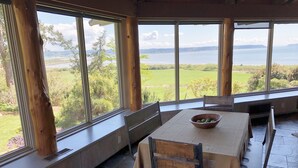 A dining area with a table and chairs, large windows, and a view of a lake and greenery. - The Alpaca Retreat - Luxury farm house with water view! (Camano Island)