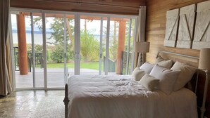 A bedroom with a large bed, a view through glass doors to a deck, and a wooden wall with a mounted artwork. - The Alpaca Retreat - Luxury farm house with water view! (Camano Island)