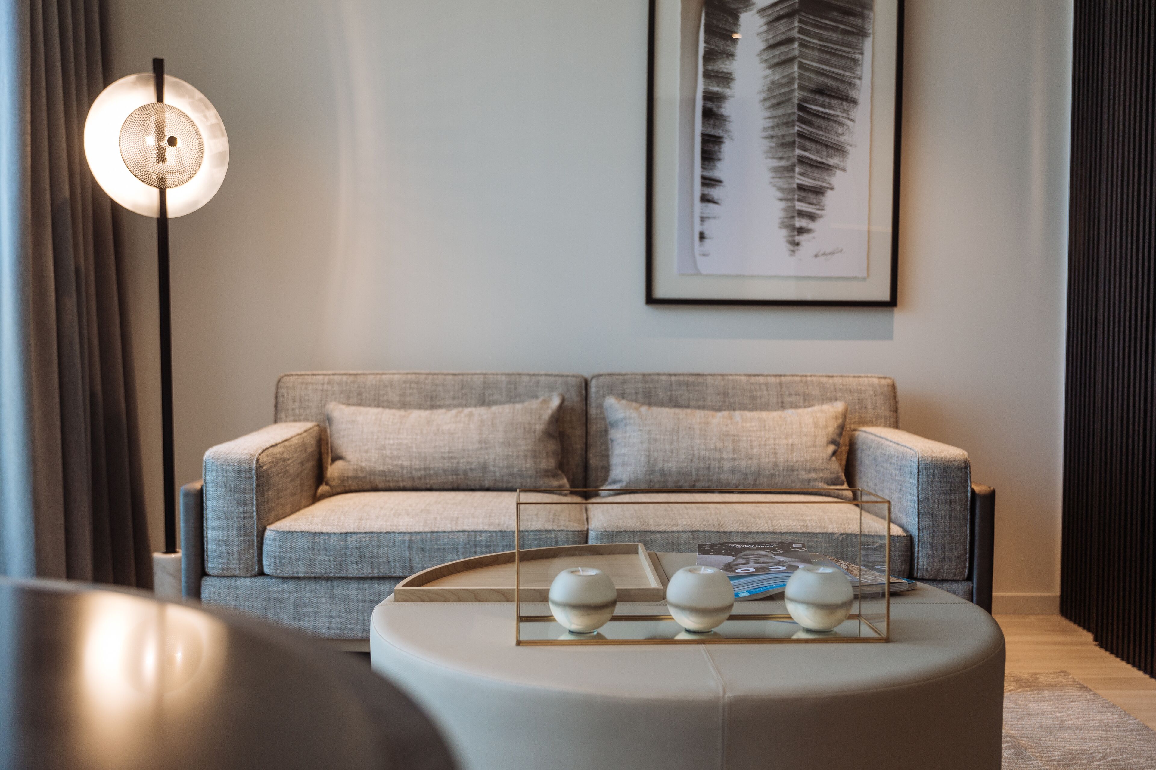 Een moderne woonkamer met een grijze bank, een ronde salontafel met decoratieve objecten, een vloerlamp en een wanddecoratie met veren.