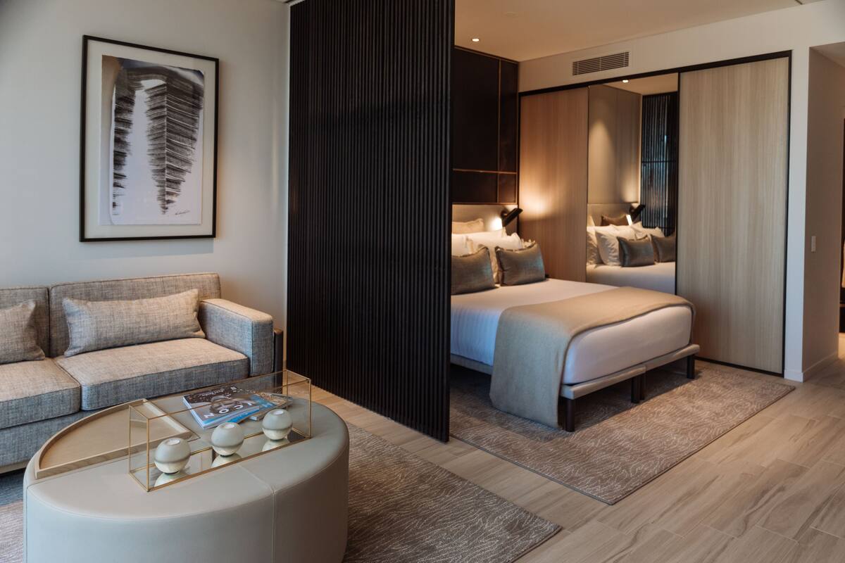 Une chambre d’hôtel moderne avec un canapé, une table basse ronde, un lit avec une tête de lit et un grand miroir.