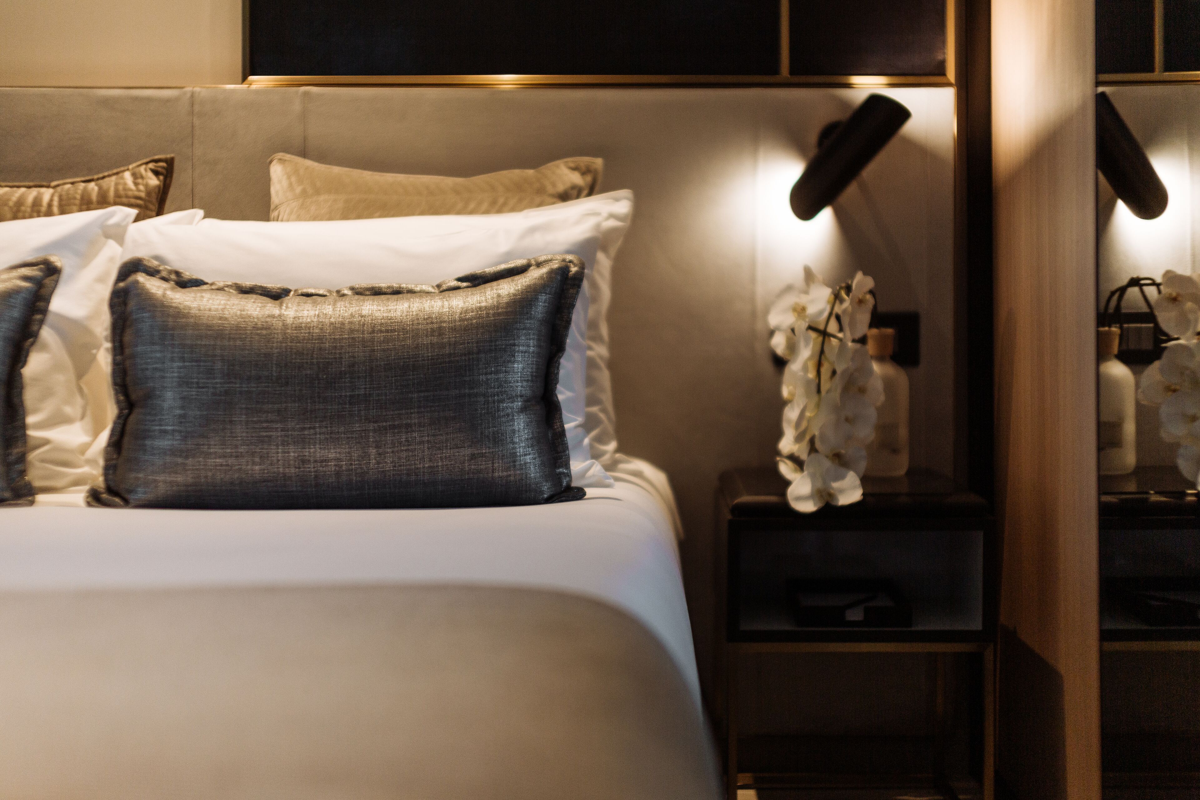 A neatly made bed with a variety of pillows, a bedside table with a lamp, and a small shelf with decorative items.