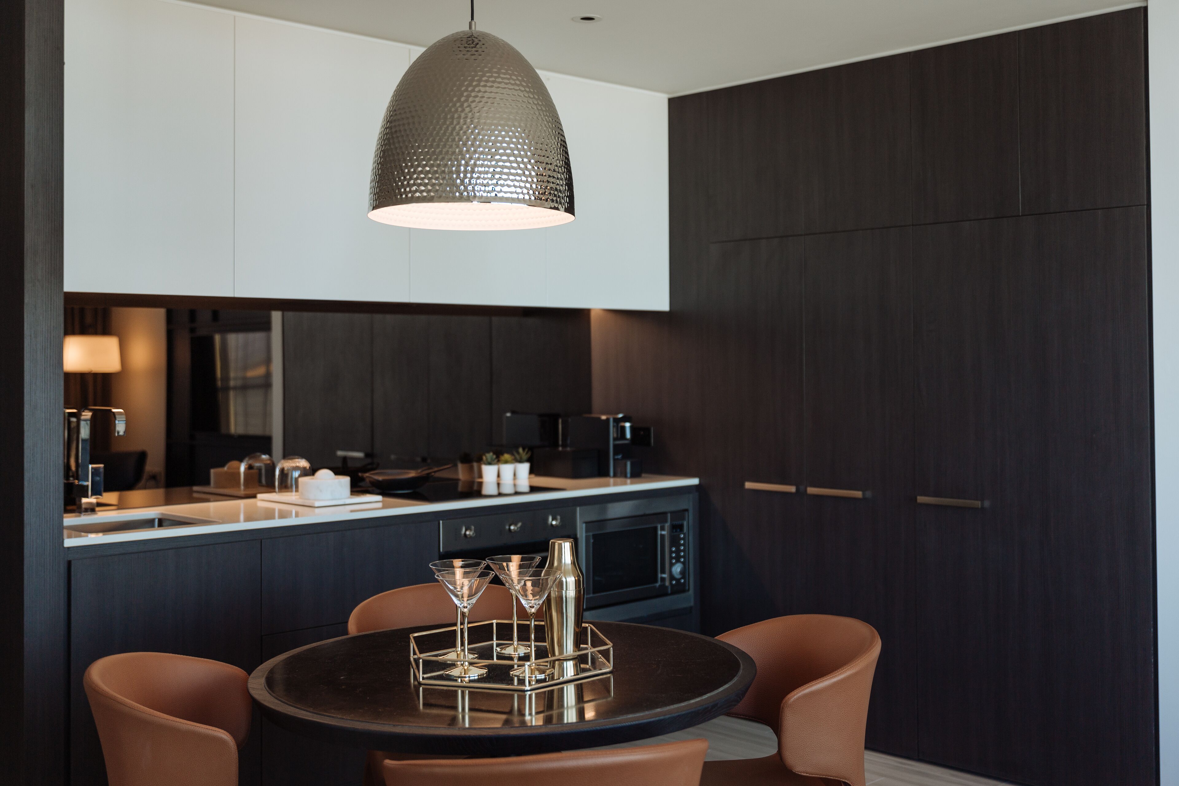 A modern kitchen with dark cabinetry, a round dining table with two chairs, and a pendant light above.