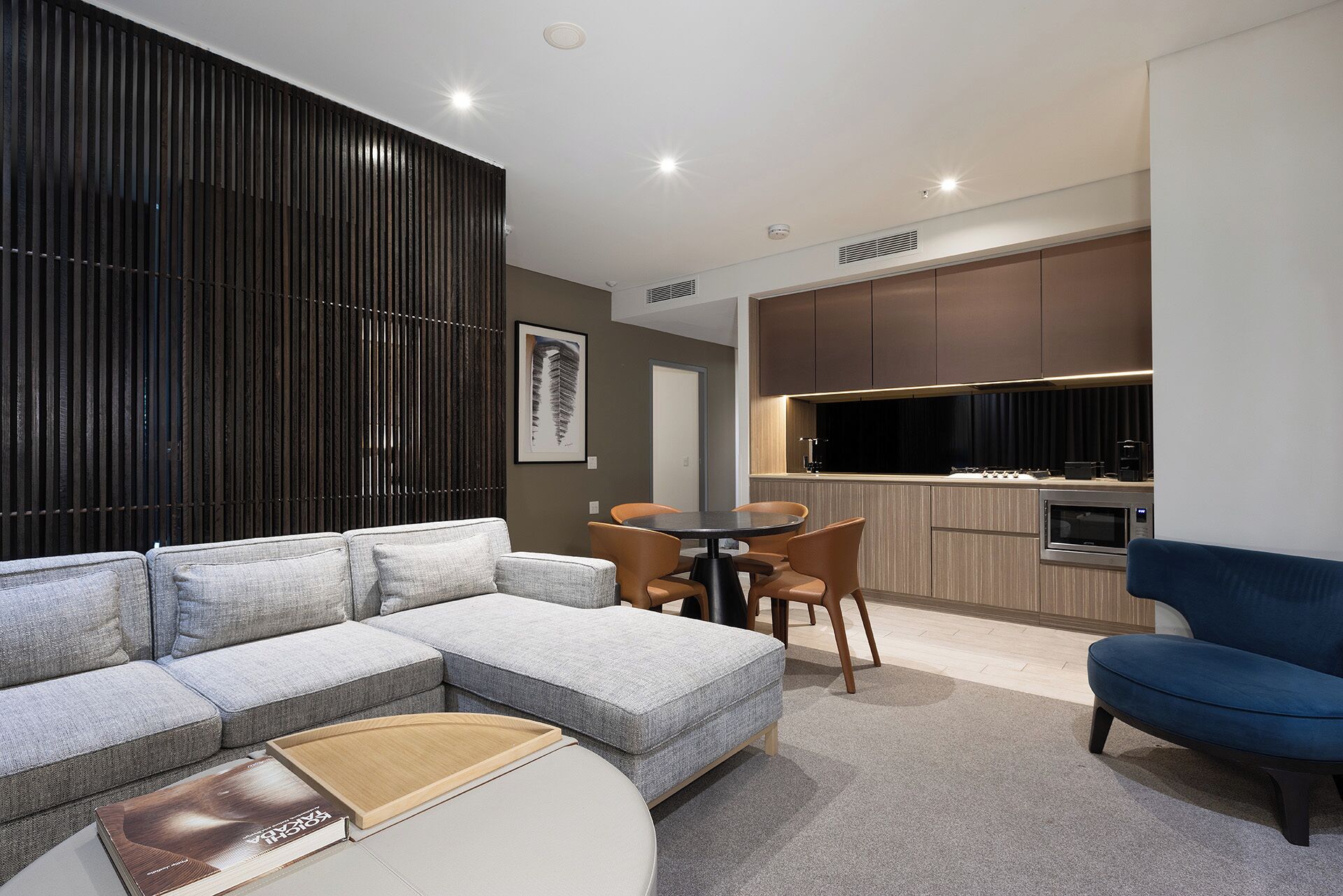 A modern living room with a grey sectional sofa, a round coffee table, and a blue armchair. There is a dining area with a small round table and chairs, and a kitchen with dark cabinets and a microwave.
