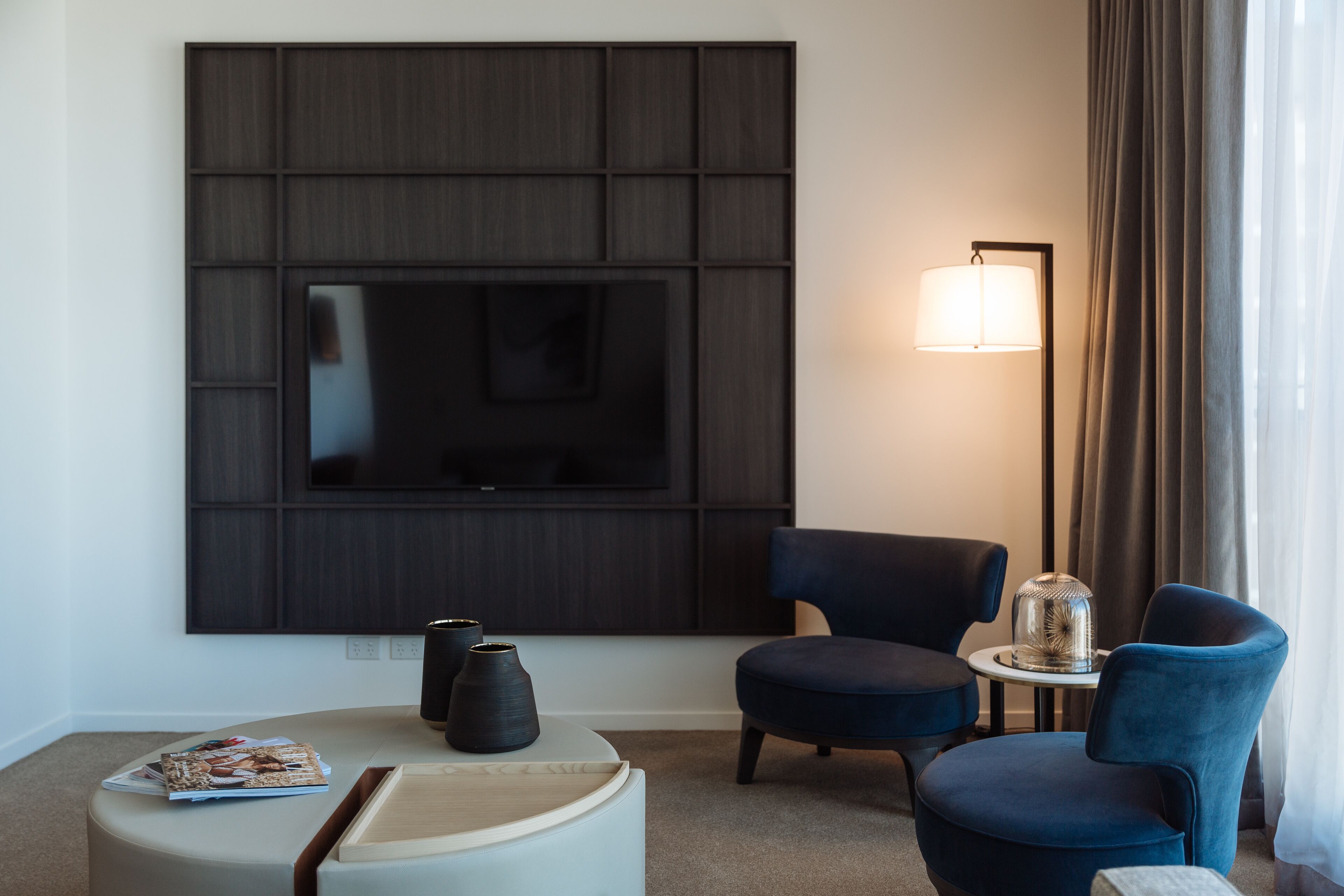A modern hotel room with a large flat-screen TV mounted on a dark panel, two blue armchairs, a small round table with a lamp, and a magazine on a white ottoman.
