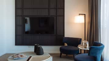 A modern hotel room with a large flat-screen TV mounted on a dark panel, two blue armchairs, a small round table with a lamp, and a magazine on a white ottoman.