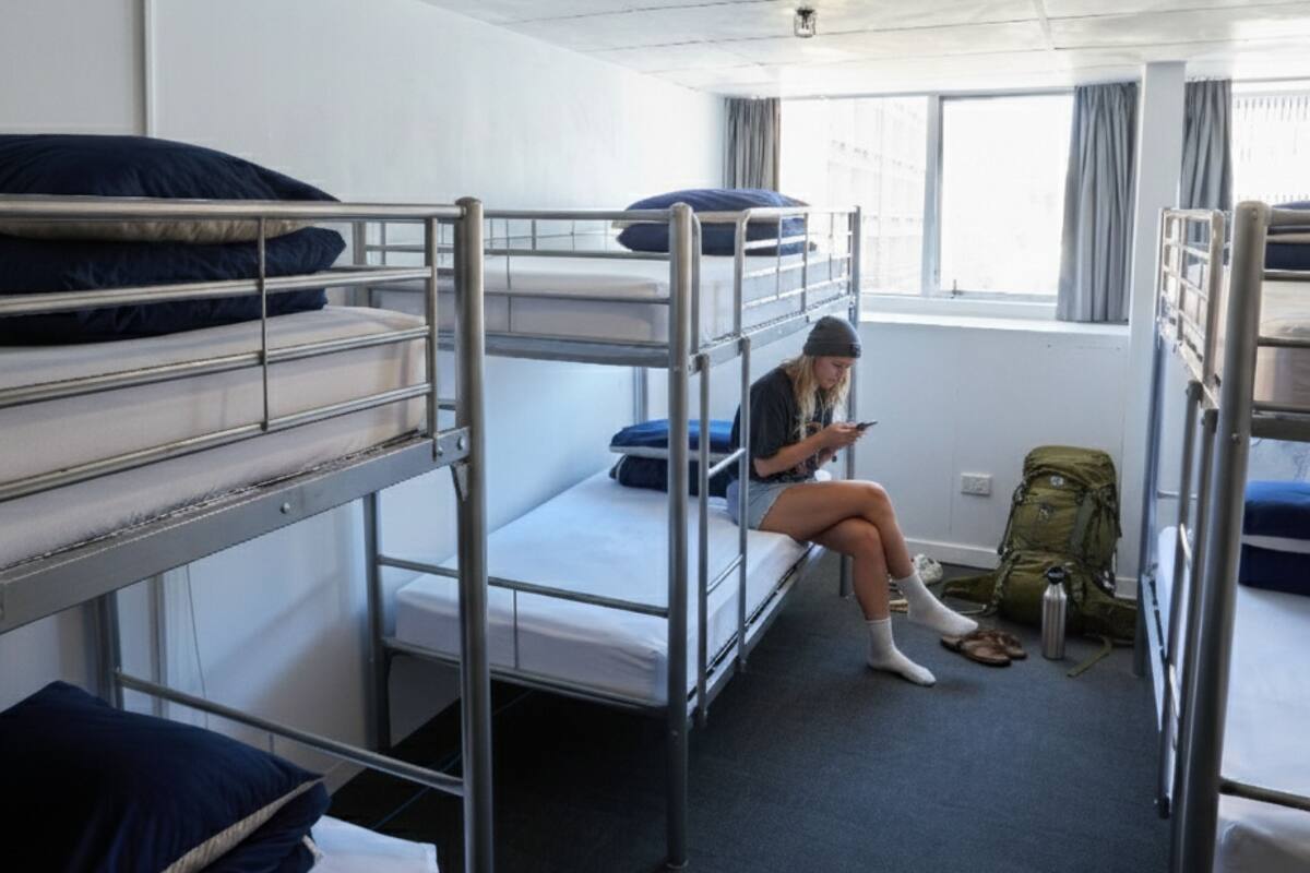 Une chambre de dortoir avec des lits superposés, une fenêtre avec des rideaux et un sac à dos posé sur le sol.