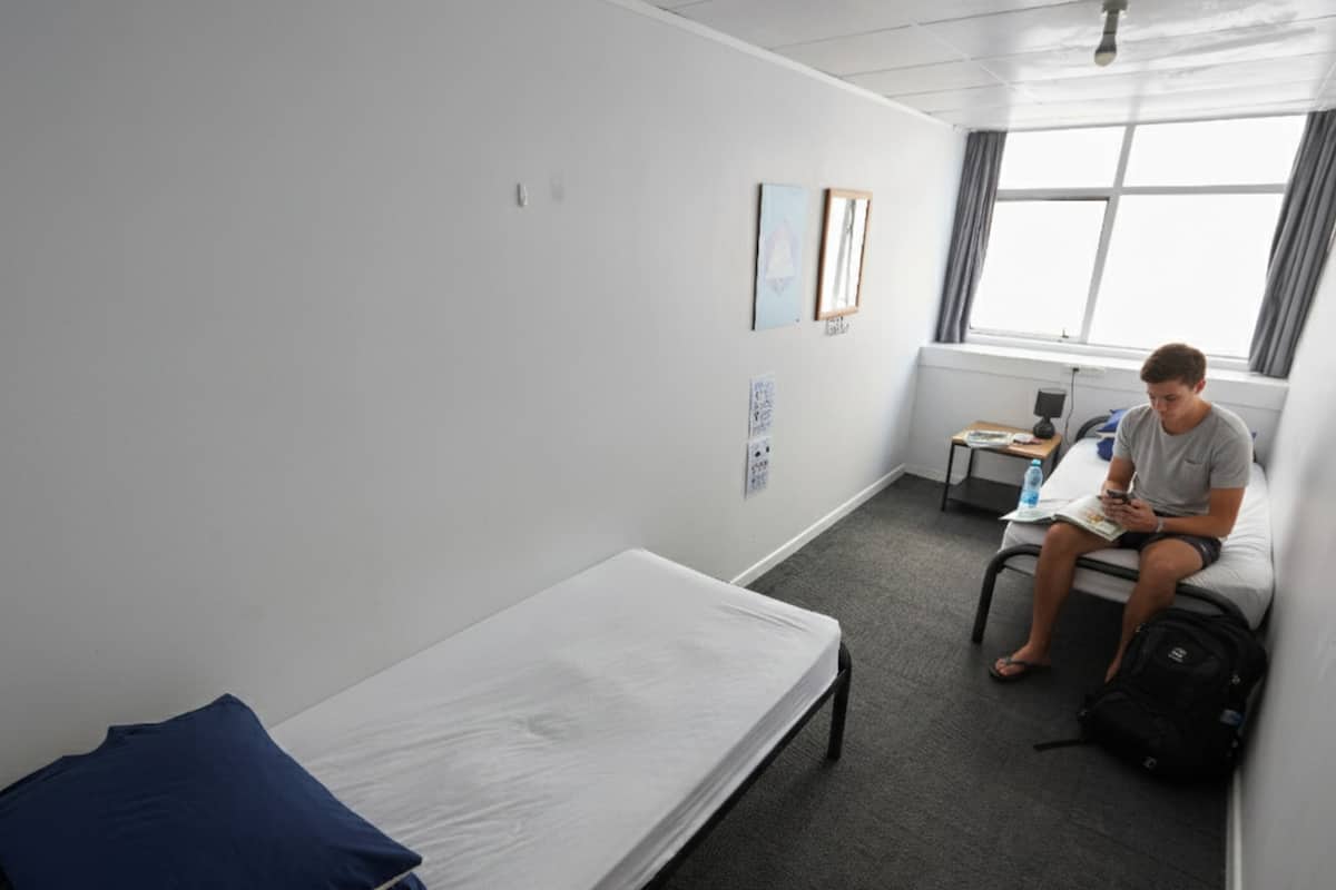 Une chambre individuelle avec un lit simple, un bureau, une chaise, une fenêtre avec des rideaux et un luminaire fixé au mur.